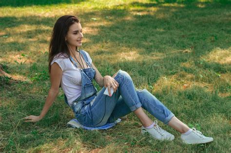 Slender Brunette Teen In Jeans Overalls Lying On The Grass In The Park