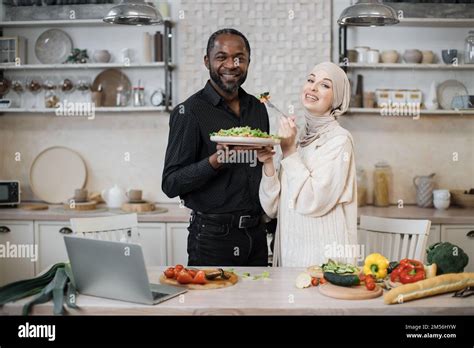 Cheerful married multinational couple using laptop while cooking
