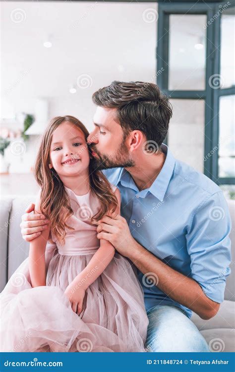 Portrait Of Father Kissing His Small Daughter Sitting On The Sofa At