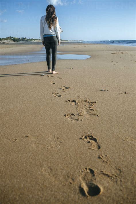 Back View Of Young Woman Walking On The Beach Leaving Her Footprints In