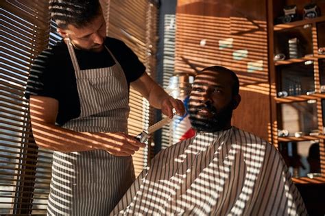 Premium Photo Male Barber In Apron Trimming Client Beard In Barbershop