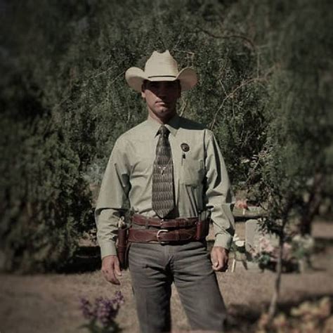 A Man Wearing A Cowboy Hat Standing In Front Of Some Trees And Bushes