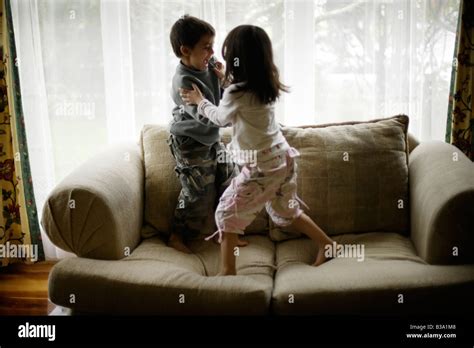 Brother Aged Six And Sister Five Wrestle On Sofa Beside Window Stock