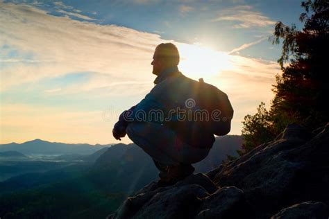 Hiker With Sporty Backpack Sit On Rocky Cliff Edge And Watching Into Misty Valley Bellow Sunny