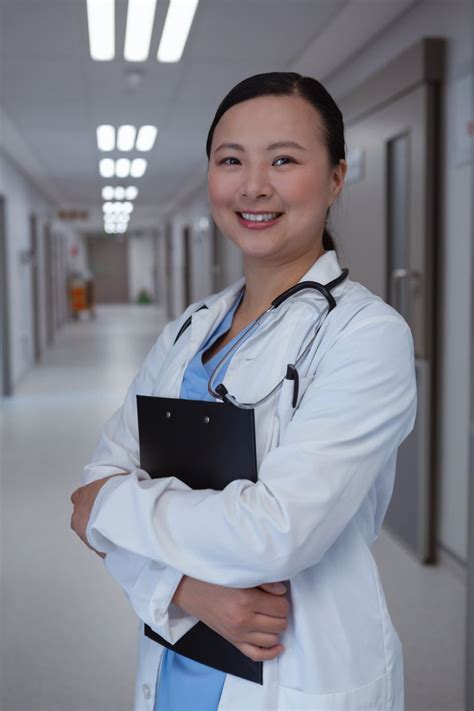 Happy Female Doctor Standing With Clipboard In Corridor At Hospital