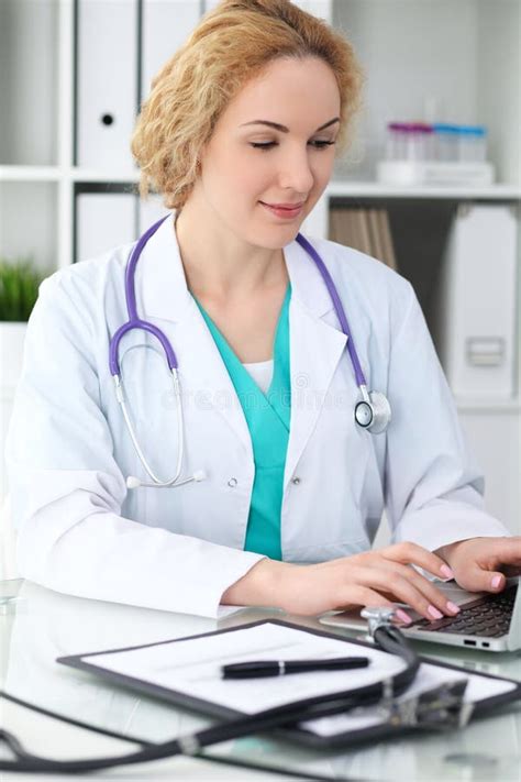 Happy Blonde Female Doctor Sitting At The Table And Typing By Laptop