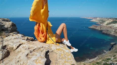 Taned Traveler Woman Sitting On Top Rock Beach With Raised Hands In Front Of Amazing Seascape