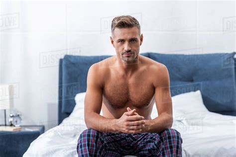 Confident Shirtless Adult Man Looking At Camera And Sitting On Bed At