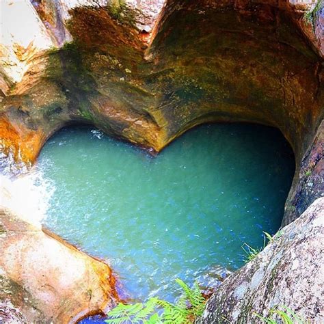 A Heart Shaped Hole In The Side Of A Mountain With Blue Water And Greenery