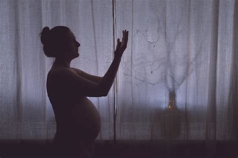 A Pregnant Woman Prays In Front Of A Night Window With Her Hands Raised In A Religious Gesture
