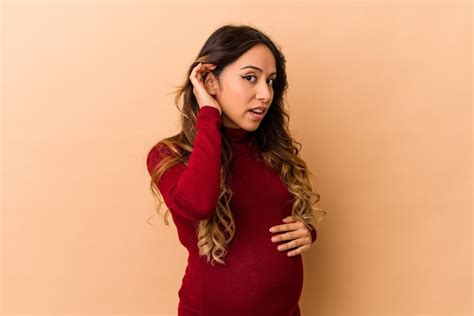 joven mujer embarazada mexicana aislada en la pared beige tratando de escuchar un chisme foto