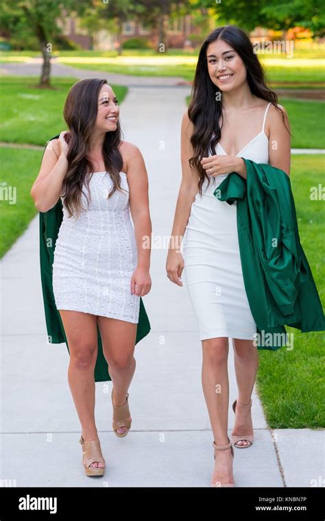 Two College Girls And Best Friends Walk On A Sidewalk While Holding Their Caps And Gowns Before