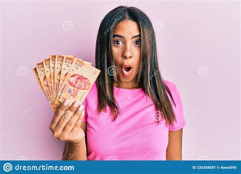 Young Latin Girl Holding Mexican Pesos Scared And Amazed With Open