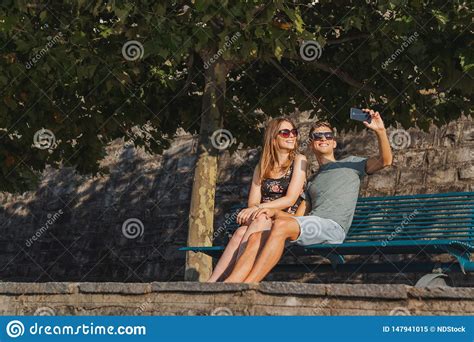 Young Couple In Love Seated On A Bench Taking A Selfie And Relaxing