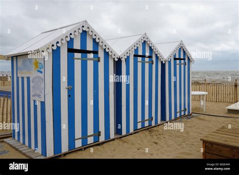 Three Wooden Striped Blue And White Beach Huts On The Sand By The Water