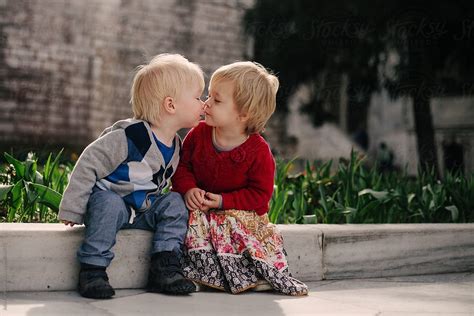 Two Small Children Sitting On A Low Wall Share A Kiss By Stocksy