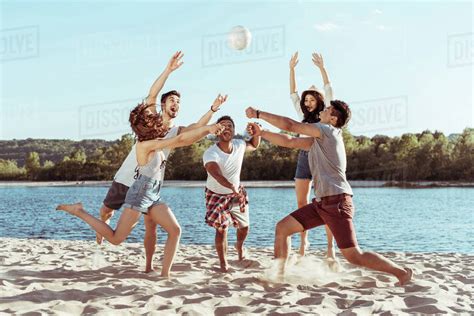 Young Smiling Friends Playing Beach Volleyball On Riverside At Daytime