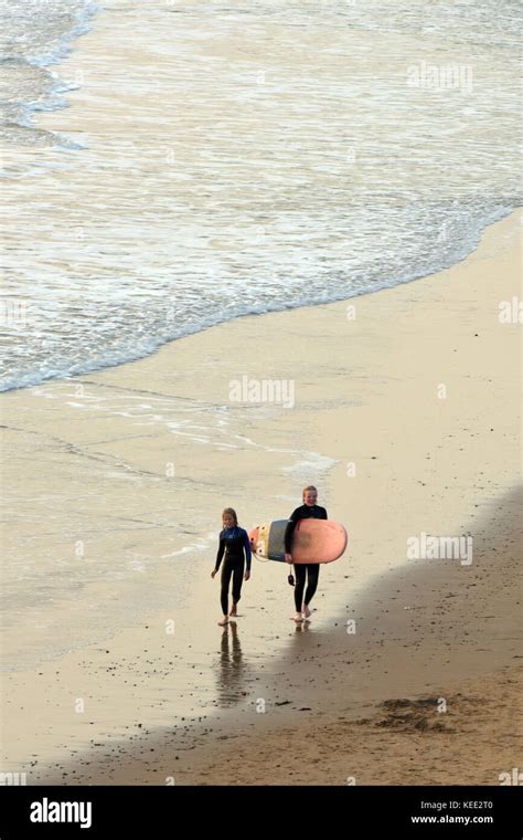 Two People Walking On A Sandy Beach At Sunset Carrying A Surfboard With The Waves And Surf At