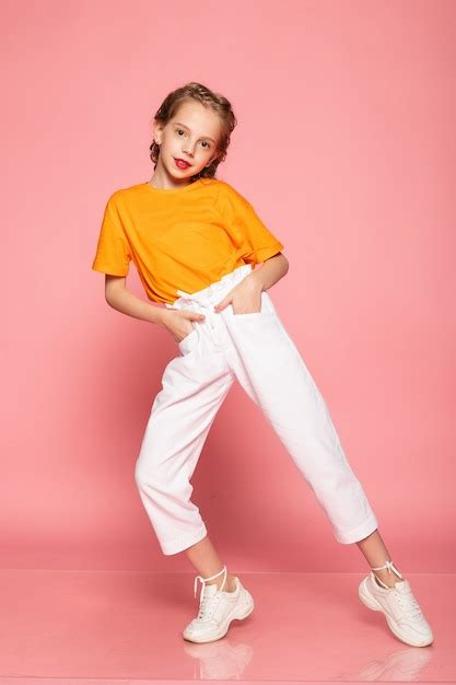 Полнометражная маленькая девочка на розовой стене студии в оранжевой футболке белых брюках и