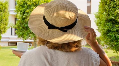 Back View Of Mature Woman In Straw Hat With White Cup Of Hot Drink Of