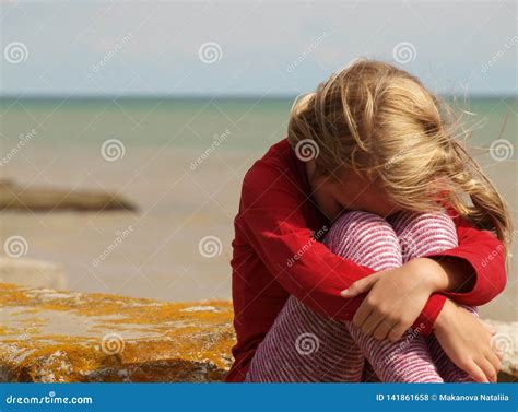 Little Girl Is Sitting Clasping Her Knees And Bowing Her Head By The