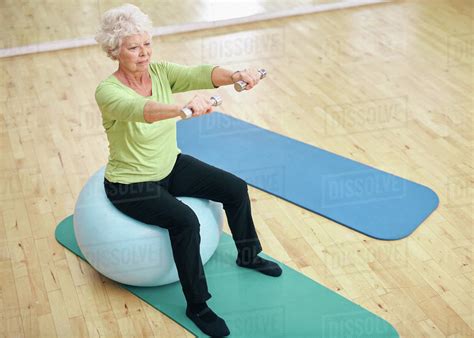 Senior Female Sitting On A Fitness Ball And Lifting Dumbbells Old