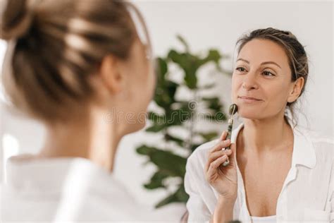 Pretty Caucasian Adult Woman Doing Face Massage With Roller Sitting In Front Of Blonde Girl