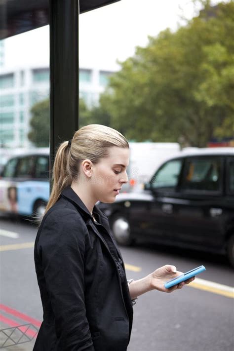 Woman Waiting At Bus Stop With Mobile Phone In London Stock Image