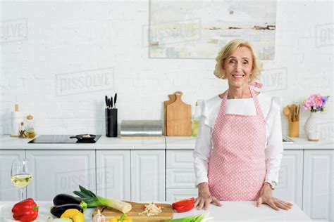 Smiling Attractive Senior Woman Standing At Kitchen Counter And Looking