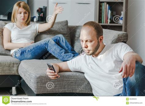 Angry Young Woman Sitting At The Sofa Talking To Her Husband While He Is Using His Cellphone In