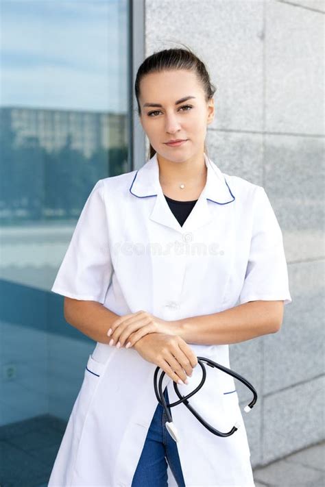 european dark haired girl doctor in white robe holds stethoscope crossed arms down outside stock