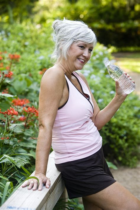 Mature Woman In Her 50s In Workout Clothes Drinking A Bottle Of Water Standing On Wooden Bridge