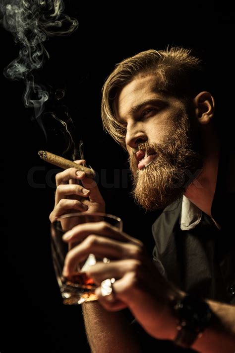 handsome bearded man holding glass of whisky and smoking cigar on black stock image colourbox