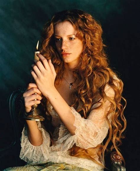A Woman With Long Red Hair Sitting On A Chair Holding A Candle In Her Hands