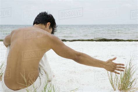 Man Sitting On The Beach Touching Dune Grass Rear View Stock Photo Dissolve