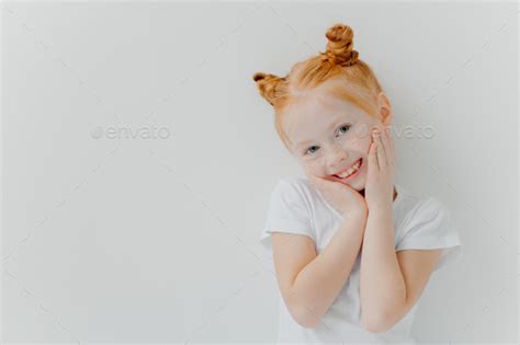 happy small redhead girl tilts head keeps hands on cheeks smiles gently at camera has good