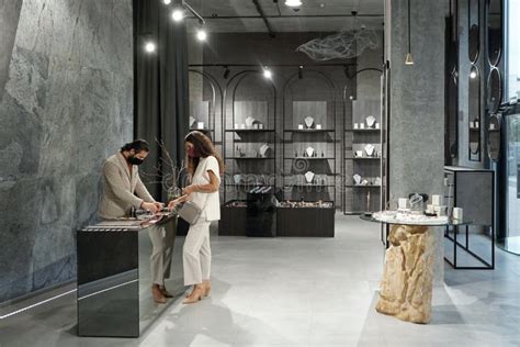 Young Female Buyer In Mask Choosing Jewelry While Male Shop Assistant Helping Her Stock Image
