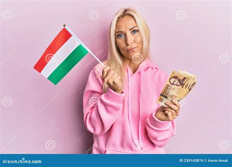 Young Blonde Woman Holding Hungary Flag And Forint Banknotes Relaxed With Serious Expression On