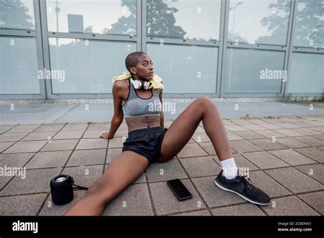 Tired Female Athlete With Shaved Head Sitting On Footpath In City Stock