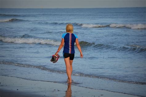 Woman In Blue And Black Shirt Holding Sneaker Walking Near Seashore