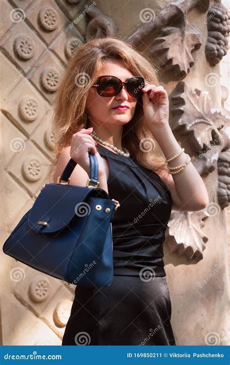 Woman In Black Dress Putting On Sunglasses And Holding Blue Bag On