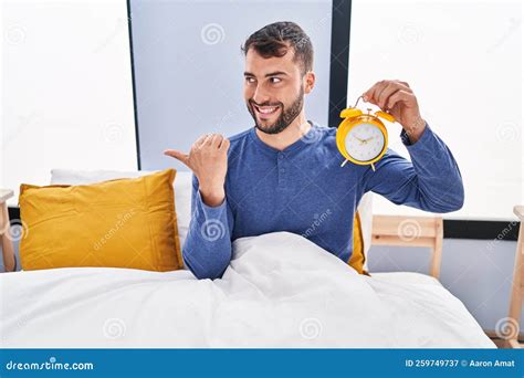 Handsome Hispanic Man In The Bed Holding Alarm Clock Pointing Thumb Up To The Side Smiling Happy