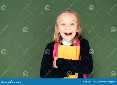 Happy Schoolgirl Looking At Camera While Holding Book And Standing Near Green Chalkboard Stock