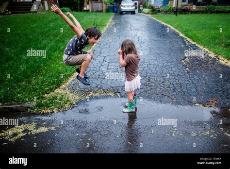 Side View Of Playful Siblings Enjoying On Wet Road During Rainy Season
