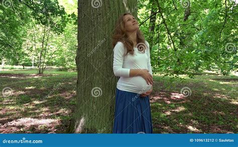 Happy Caucasian Pregnant Girl Stroking Her Belly Standing Near Big Tree Trunk Stock Footage