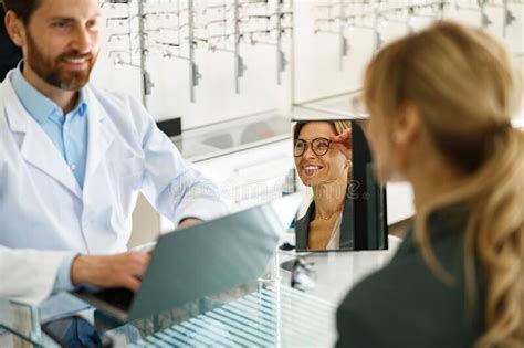 male optometrist helping woman to choose glasses in optical store showing to her mirror stock