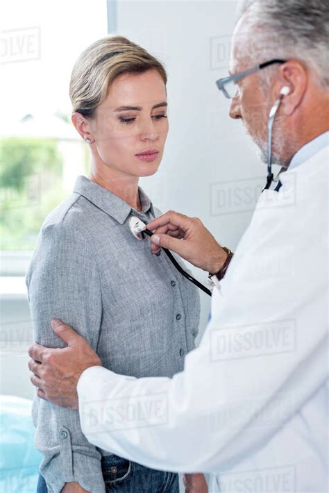 Mature Male Doctor Examining Female Patient By Stethoscope In Hospital