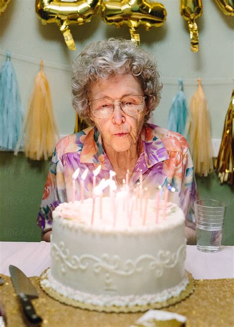 Elderly Woman Blowing Out Birthday Candles On Cake By Stocksy