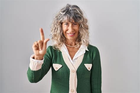 흰색 배경 위에 서서 손가락 1번을 가리키며 자신감 있고 행복한 미소를 짓고 있는 중년 여성 무료 사진