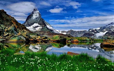 Sharp Mountain Peaks Of Rock Snow Lake Meadow With Green Grass Blue Sky With White Clouds